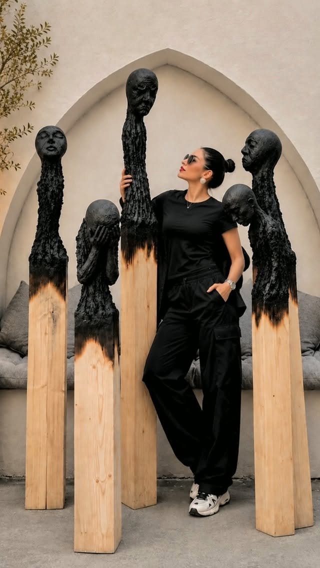 Art in Wood: 8 Ideas, From Knot Jewelry to Totem Pillars 7 Woman standing next to tall wooden pillars carved into black artistic human heads.