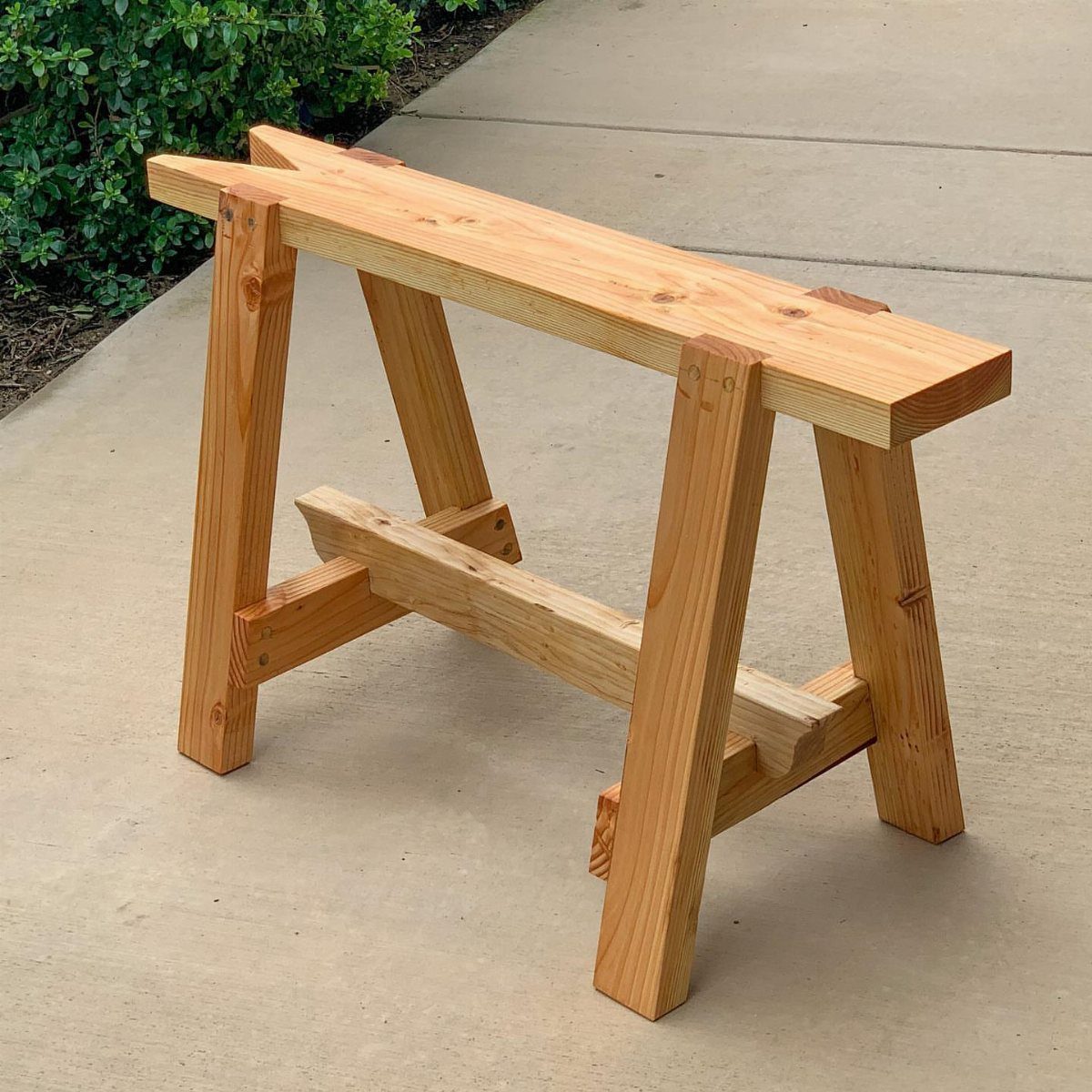 13 Carpentry Tools and Equipment for Every Workshop 8 Wooden sawhorse made of light pine lumber standing on a concrete walkway.