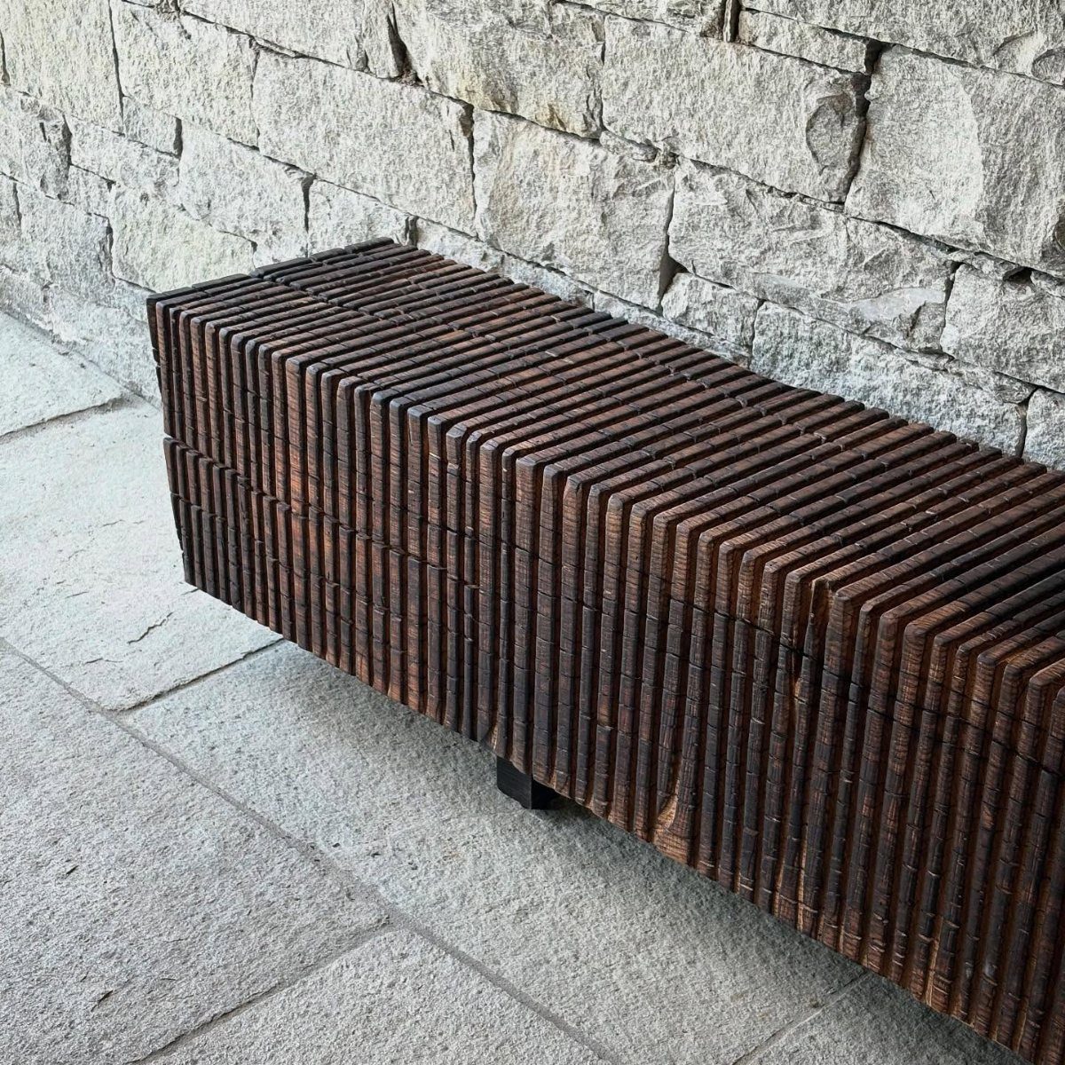 Charred wood bench with a ribbed texture against a grey stone wall.