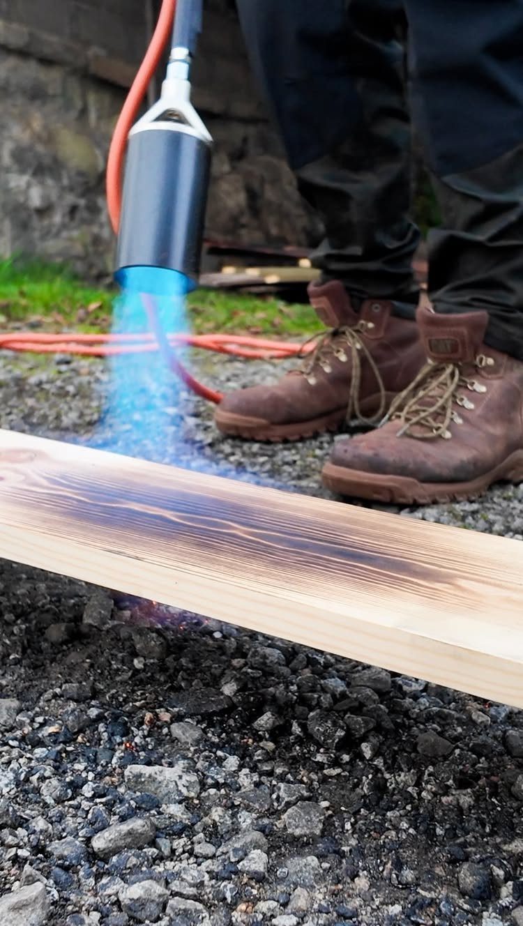Propane torch applying a blue flame to char a wooden plank on gravel.