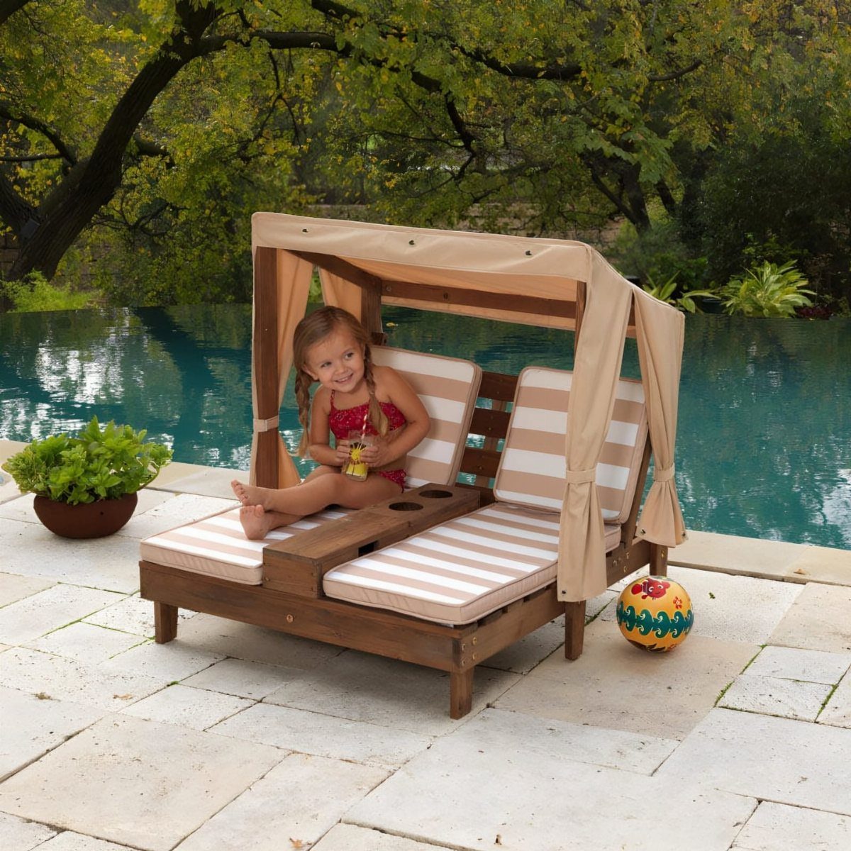 Child sitting on a wooden double chaise lounge with a canopy by a pool.