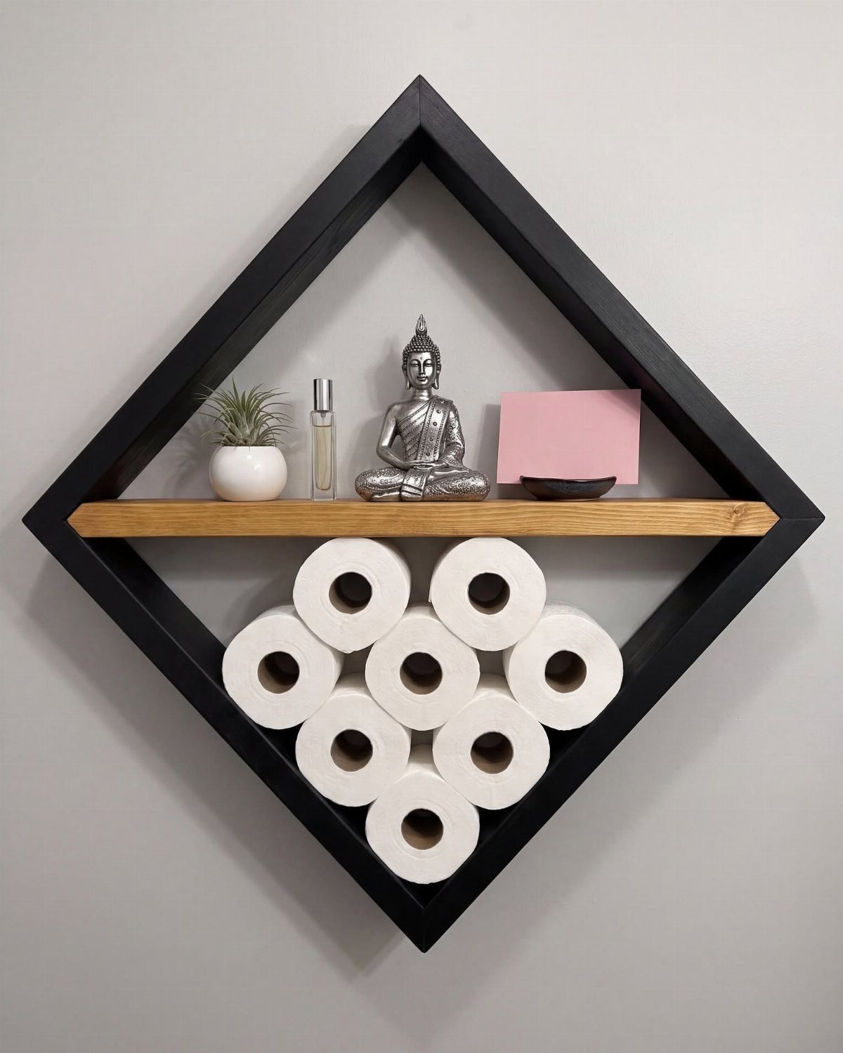 Diamond-shaped black wooden shelf holding toilet paper rolls and decorative items on a wall