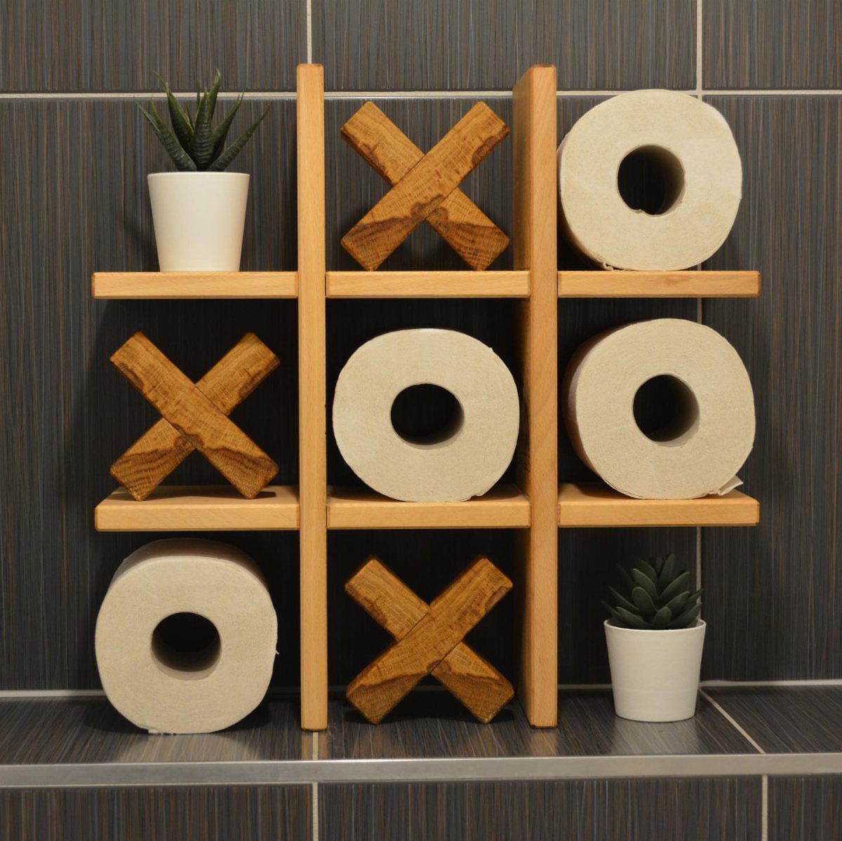 Wooden tic-tac-toe shelf holding toilet paper rolls and small potted succulents.