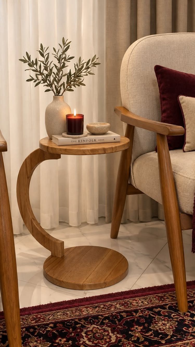 C-shaped wooden side table with a vase, candle, and book next to an armchair.