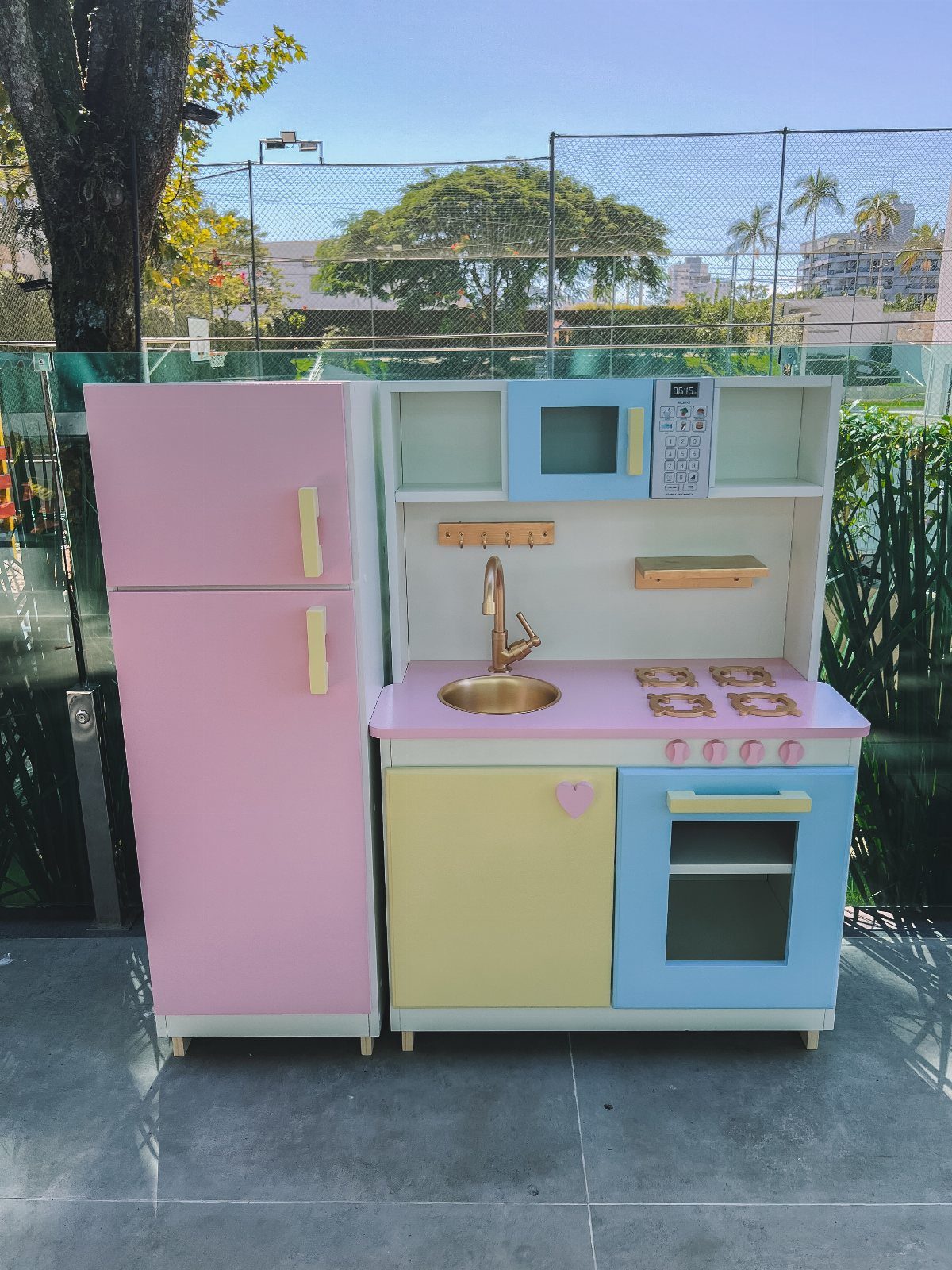 Wooden kids play kitchen with pink refrigerator, yellow cabinet, and blue oven outdoors.