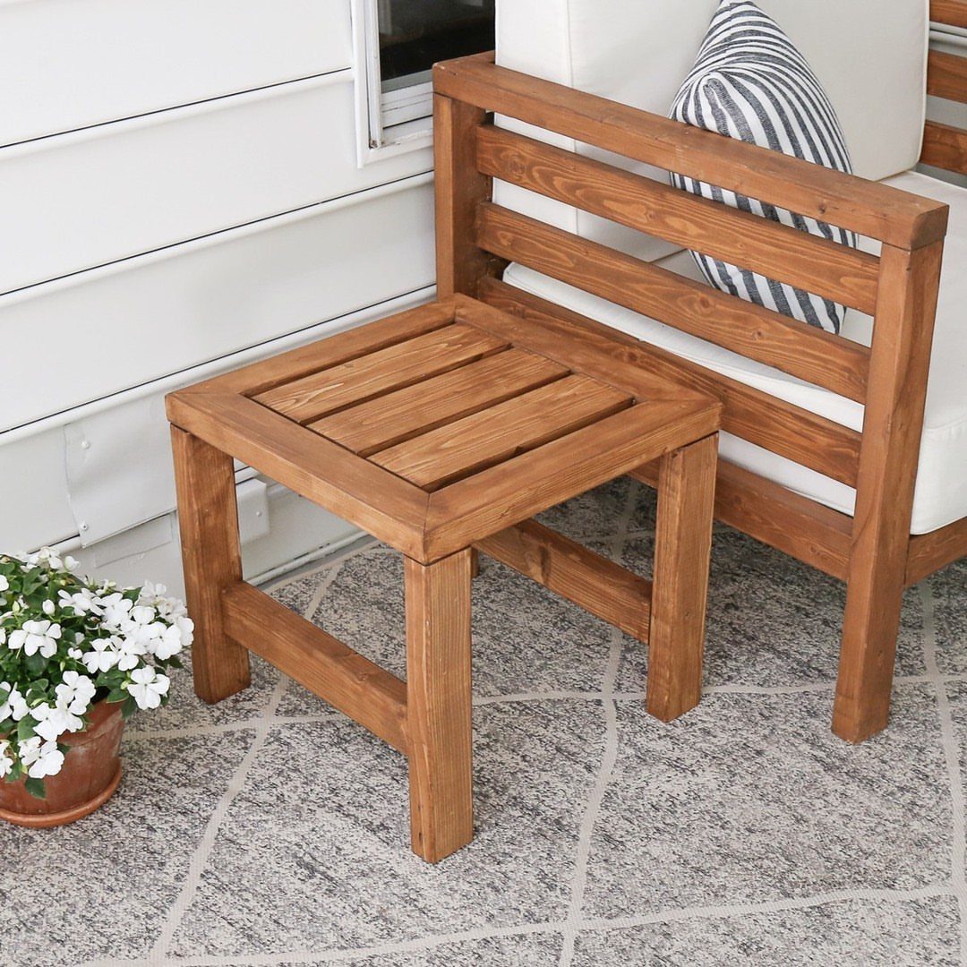 Wooden outdoor side table next to a cushioned wooden chair on a stone patio.