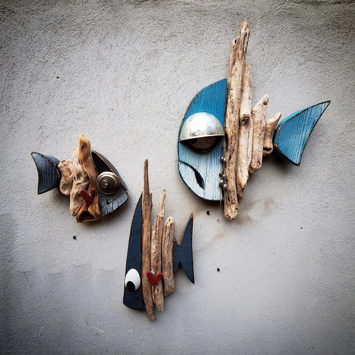 Three fish sculptures crafted from blue wood and driftwood on a grey wall.