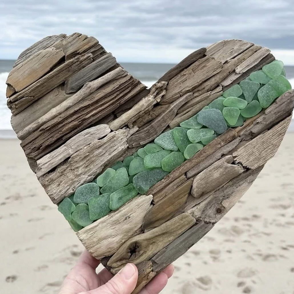 Heart-shaped driftwood sculpture with a diagonal line of green sea glass on a beach.