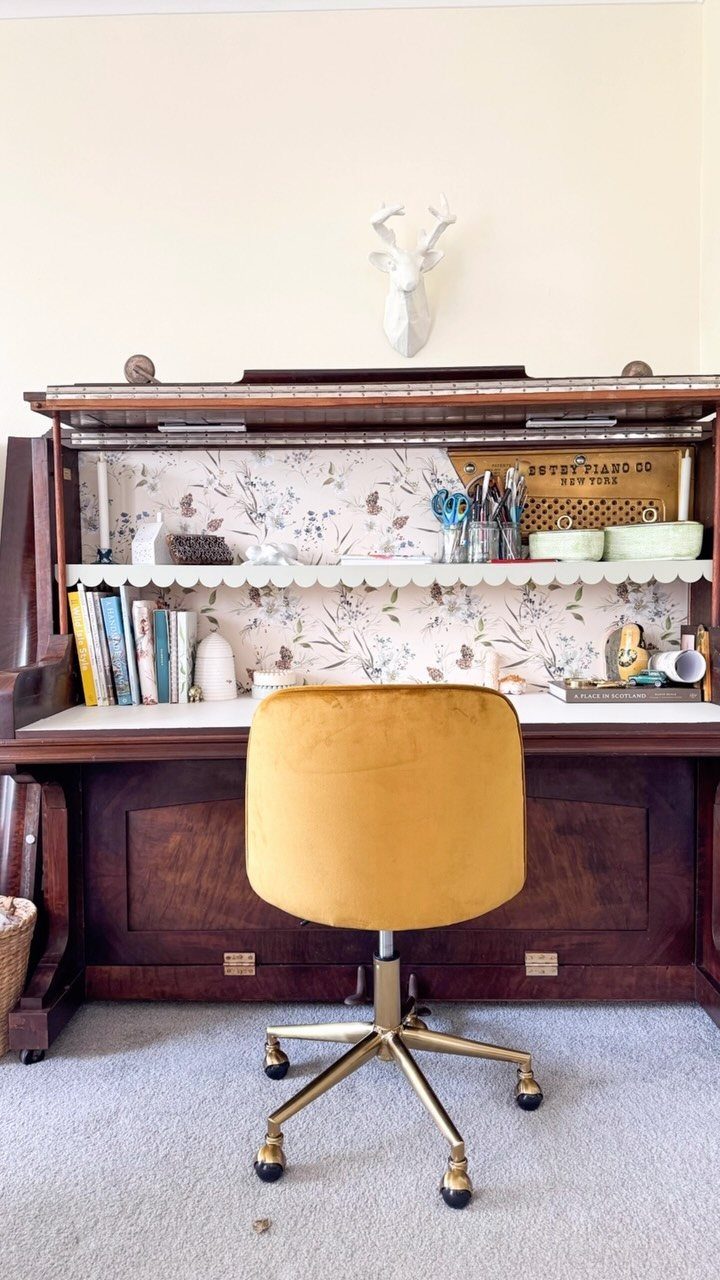 Converted upright piano desk with floral wallpaper and a yellow velvet swivel chair.