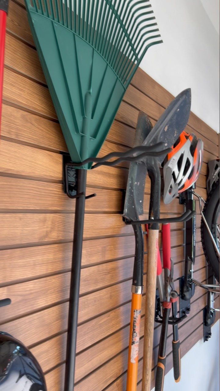 Green rake and shovels hanging on a brown wooden slatwall storage system.