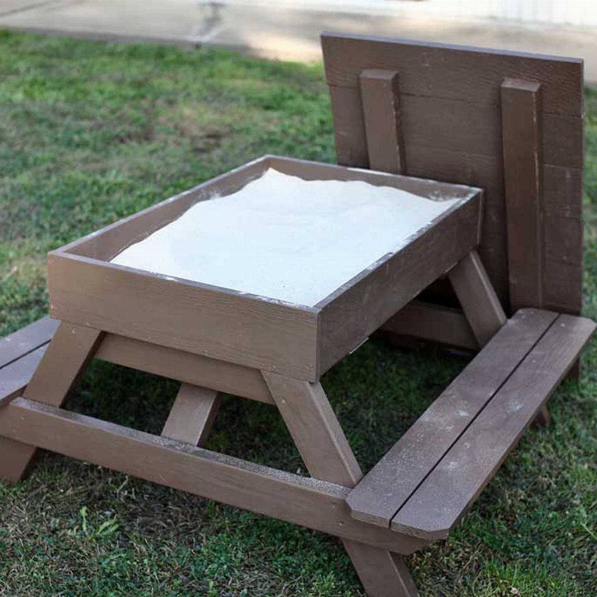 Brown wooden sandbox with built-in benches and a backrest on a grassy lawn.