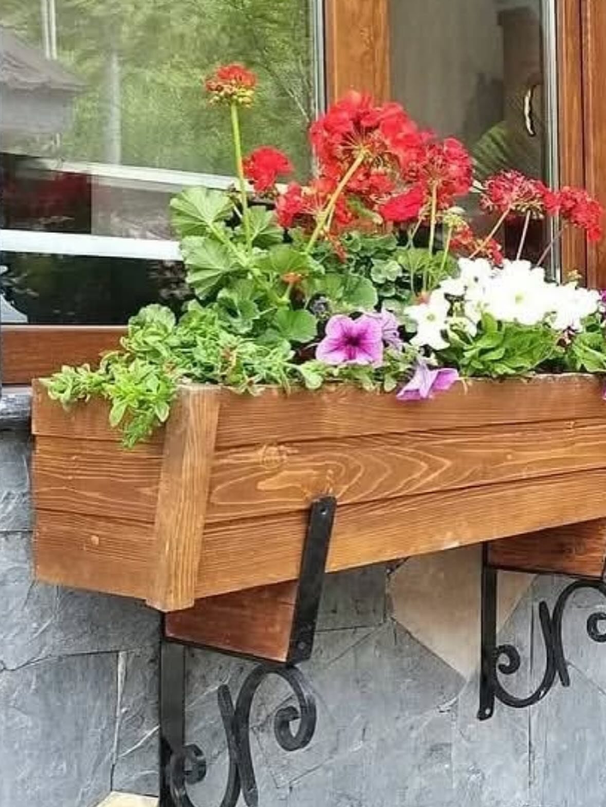 Wooden window flower box with colorful blooms held by black decorative metal brackets.