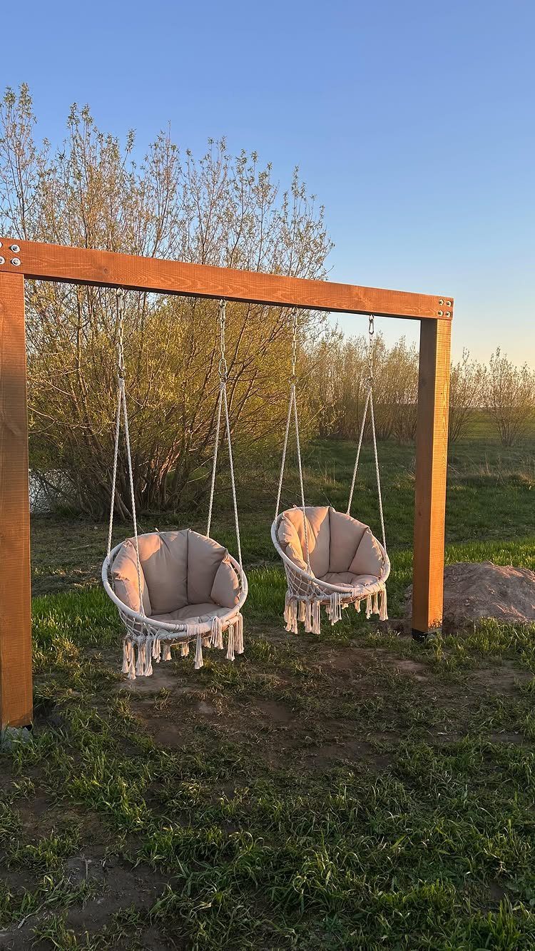 Two beige macrame hanging chairs suspended from a wooden beam in a garden.