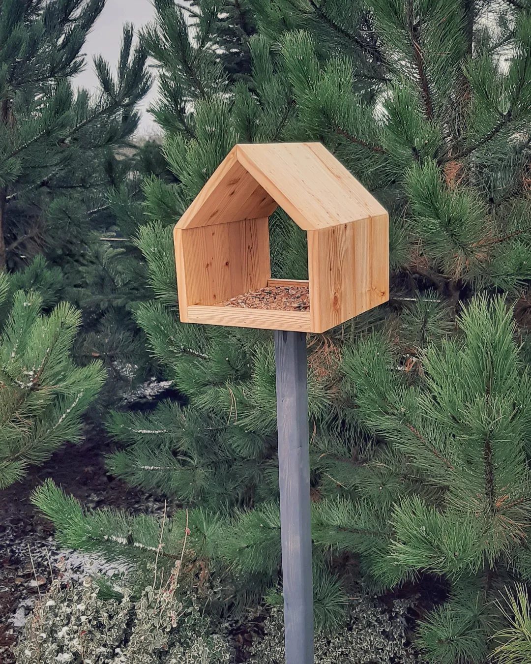 32 Mini Wood Projects for Beginners and Scrap Wood 20 Wooden bird feeder with a peaked roof mounted on a pole among pine trees.