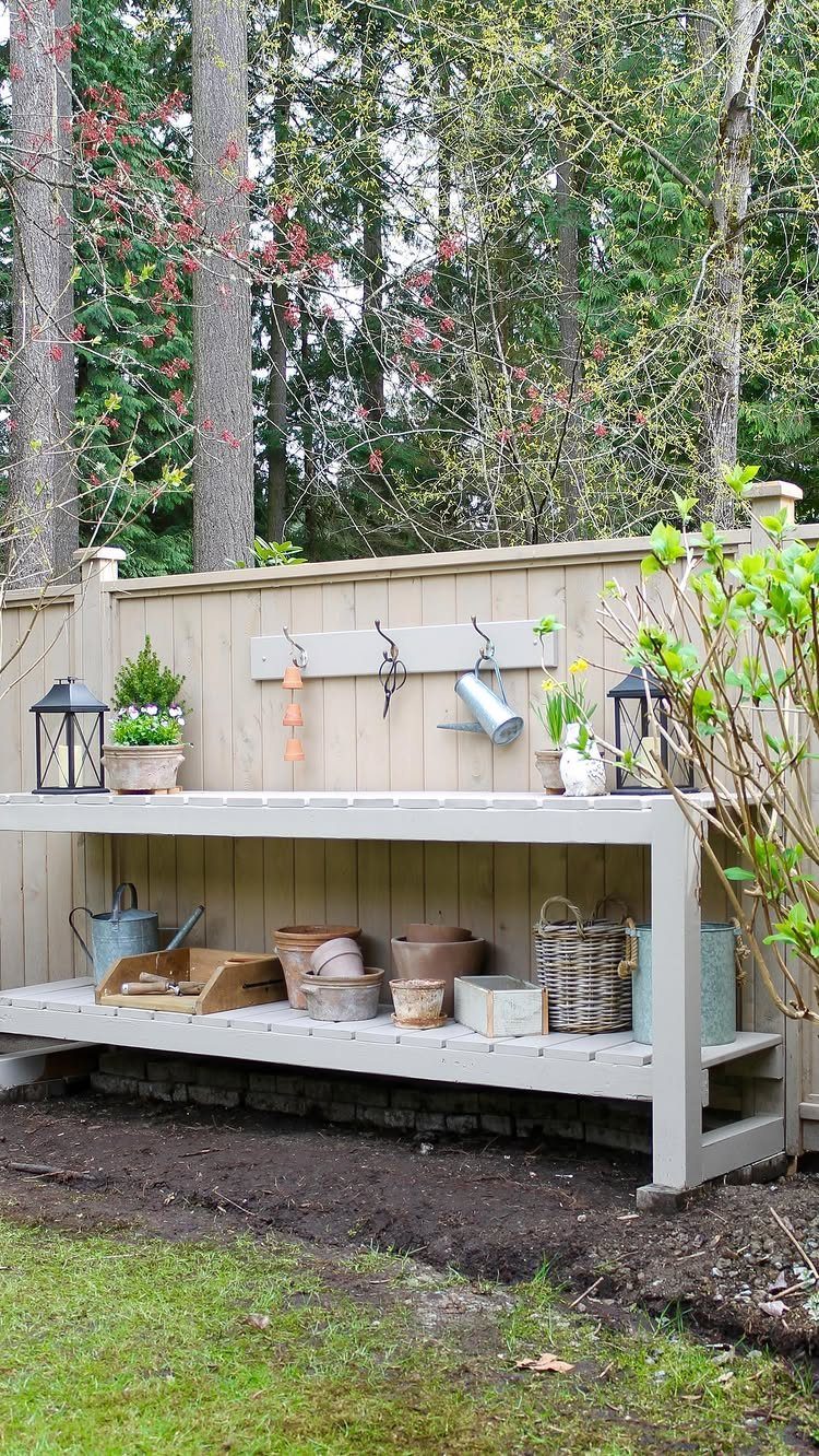 Light grey wooden potting bench with gardening tools against a tan wooden fence.