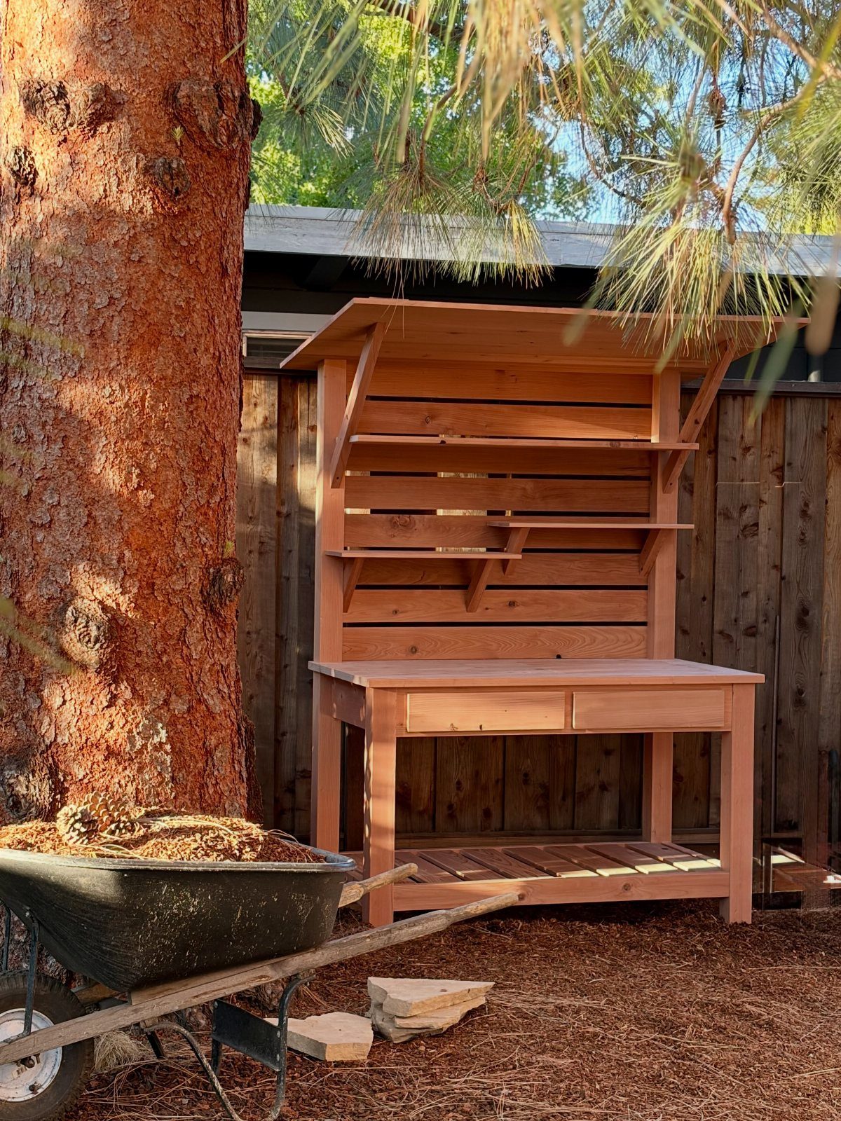 Wooden potting bench with shelves and roof standing outdoors next to a tree.