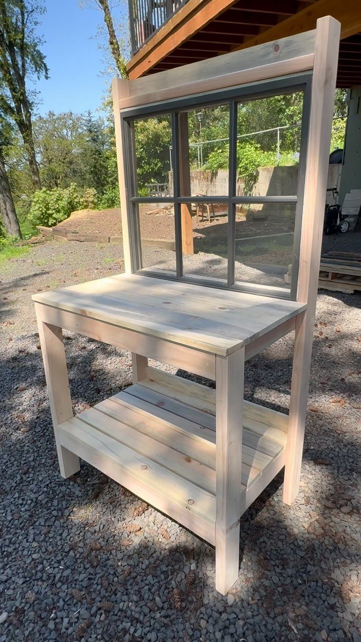 Wooden potting bench with a lower shelf and a window frame backsplash outdoors.