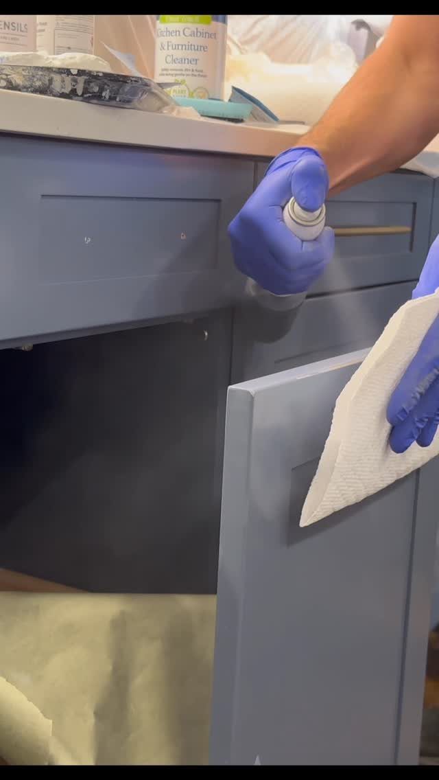 Person in blue gloves cleaning a grey-blue wooden cabinet with spray and cloth.