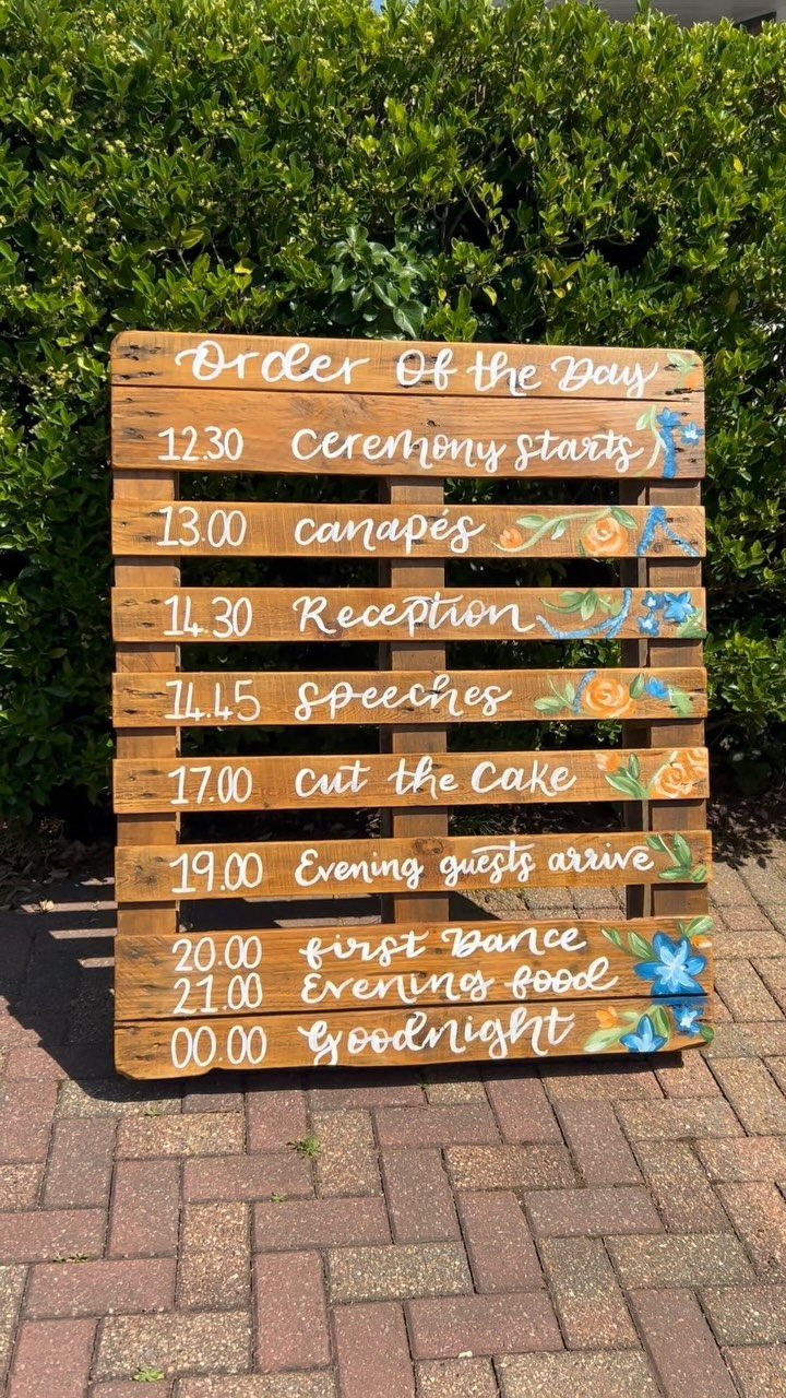 Rustic pallet wood sign with wedding schedule and floral paintings against a green hedge.