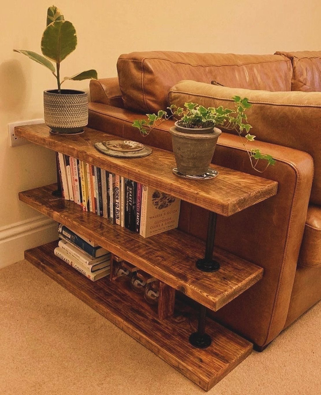 Three tiered stained wood shelves with books and plants next to a leather sofa.
