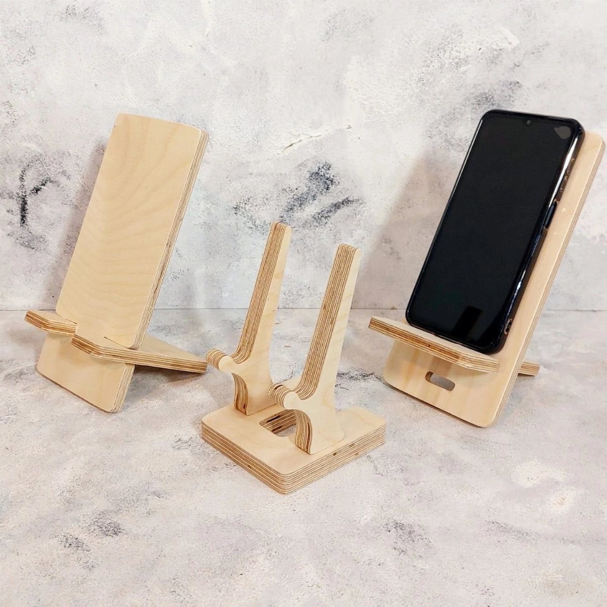 13 Small Wood Craft Ideas for Beginners and Scrap Wood 11 Three light plywood phone stands, one holding a black smartphone, on grey background.