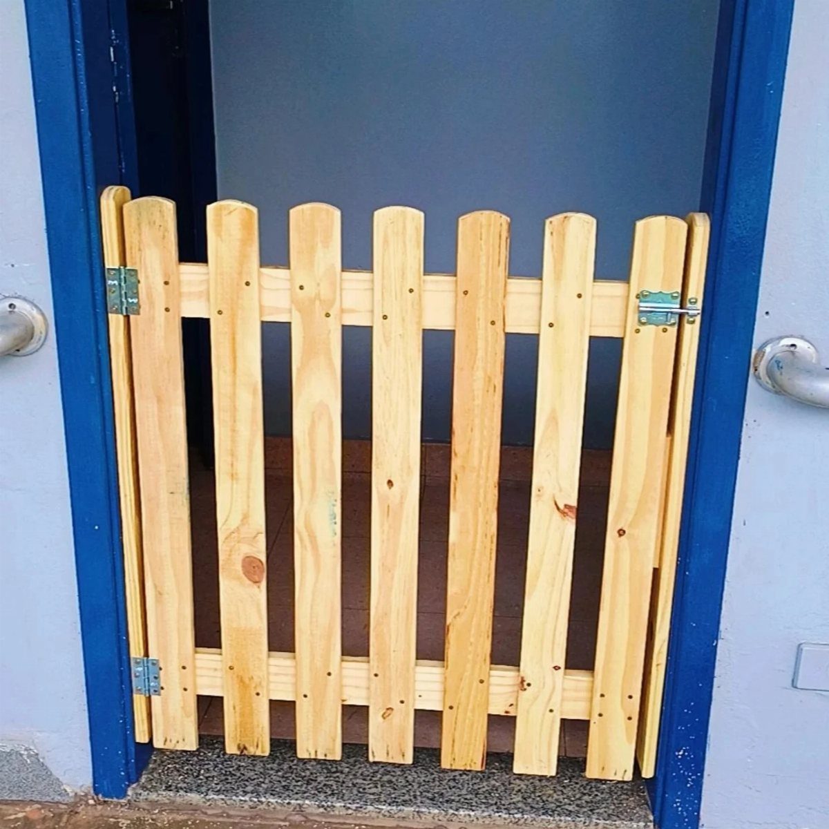 Wooden picket pet gate installed in a blue doorway with silver metal hinges.
