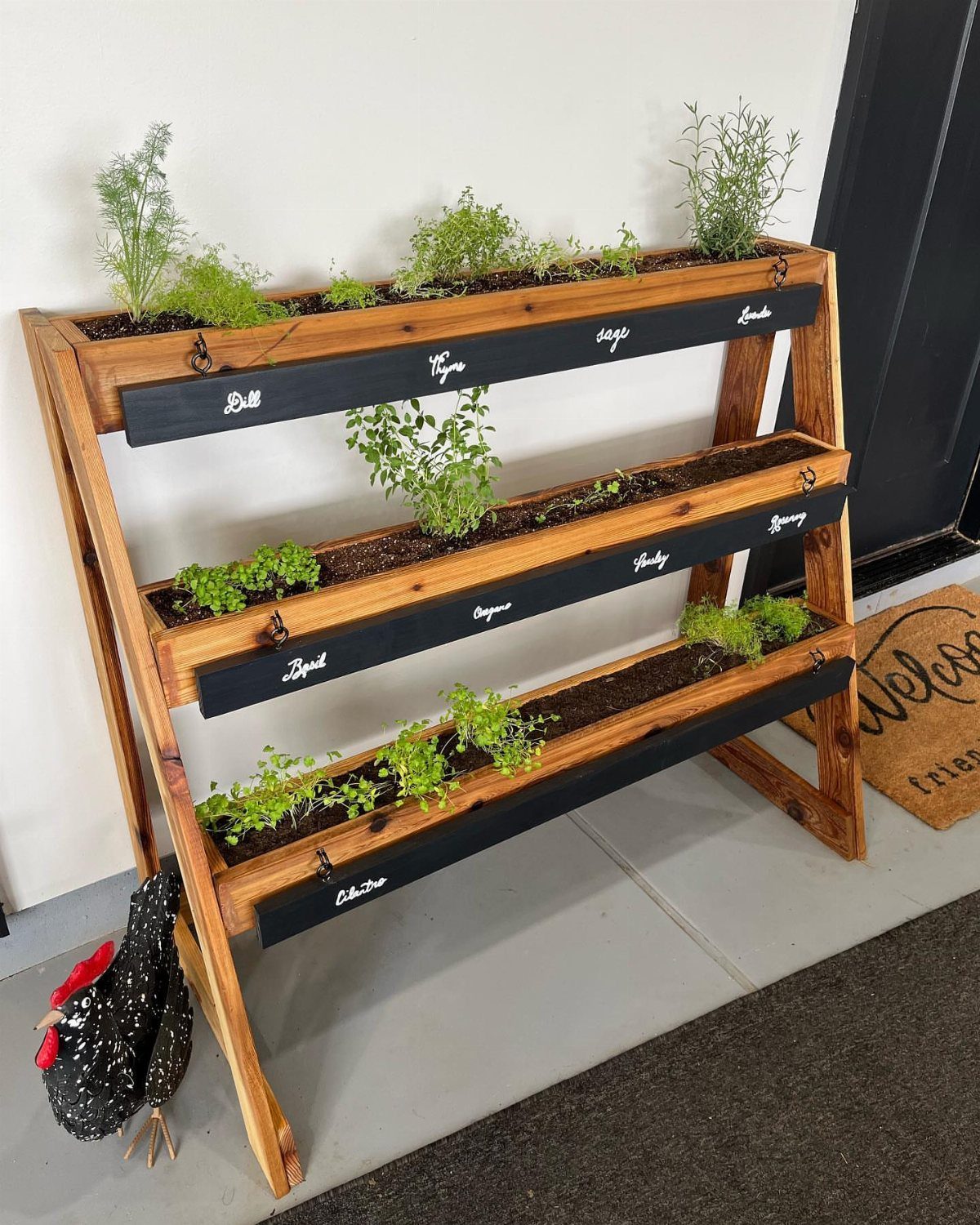 Three-tiered wooden herb planter with black labels holding various green plants indoors.