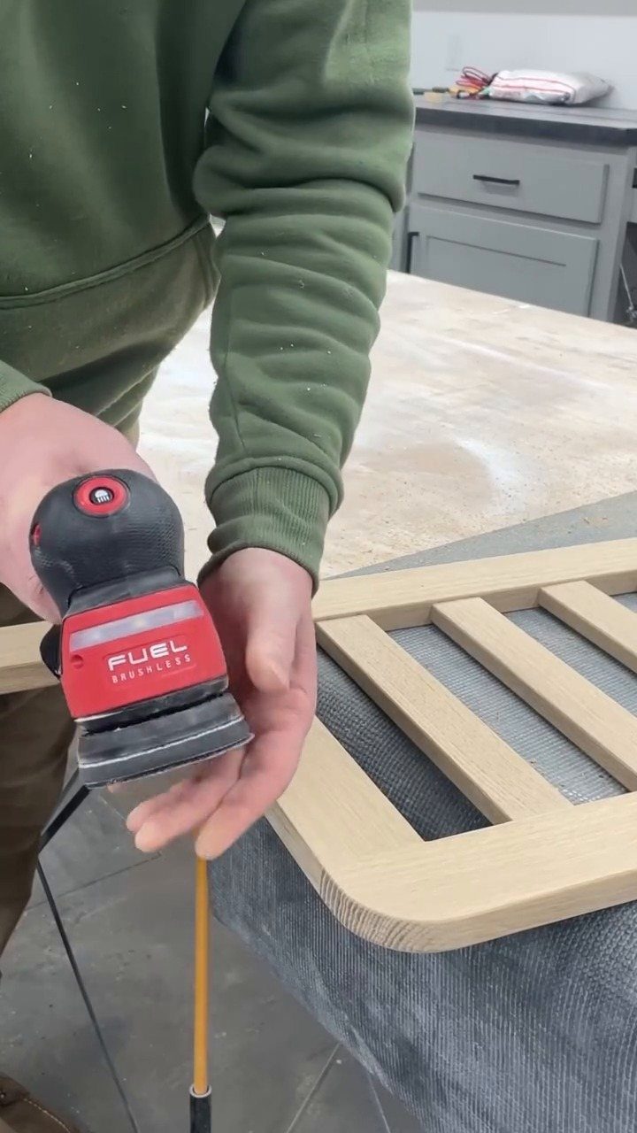 Person using a red power sander on a light-colored wooden frame in a workshop.