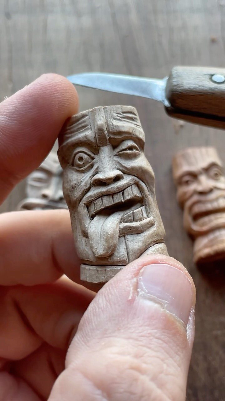 Carved wooden face with a protruding tongue being worked on with a whittling knife.