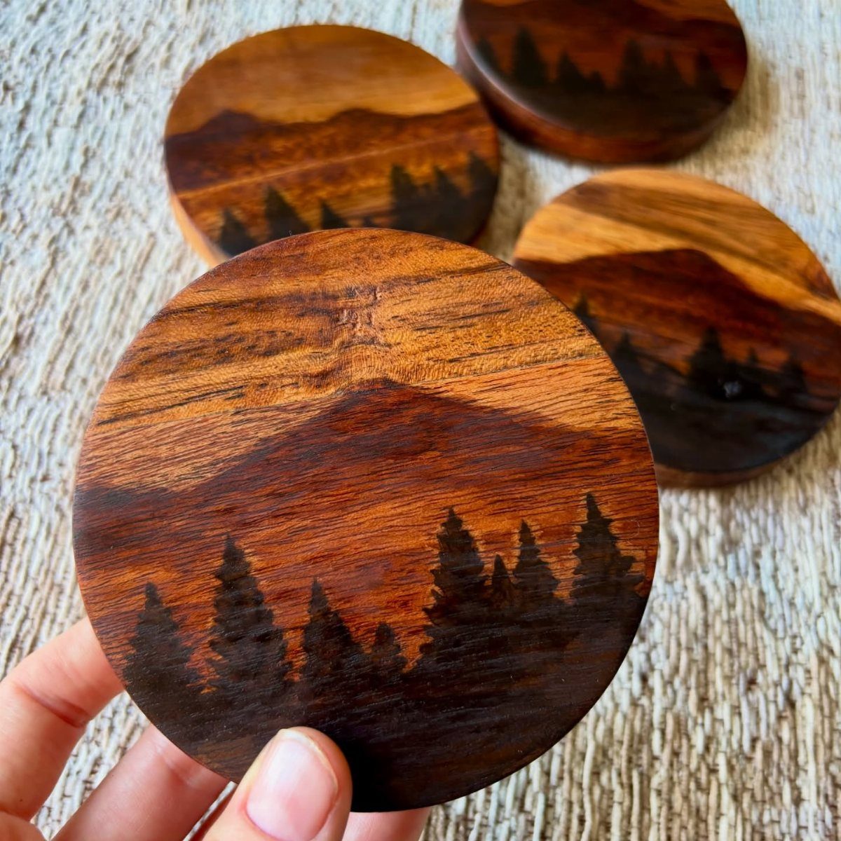 Circular wooden coasters with wood-burned mountain and forest landscapes on a textured background.
