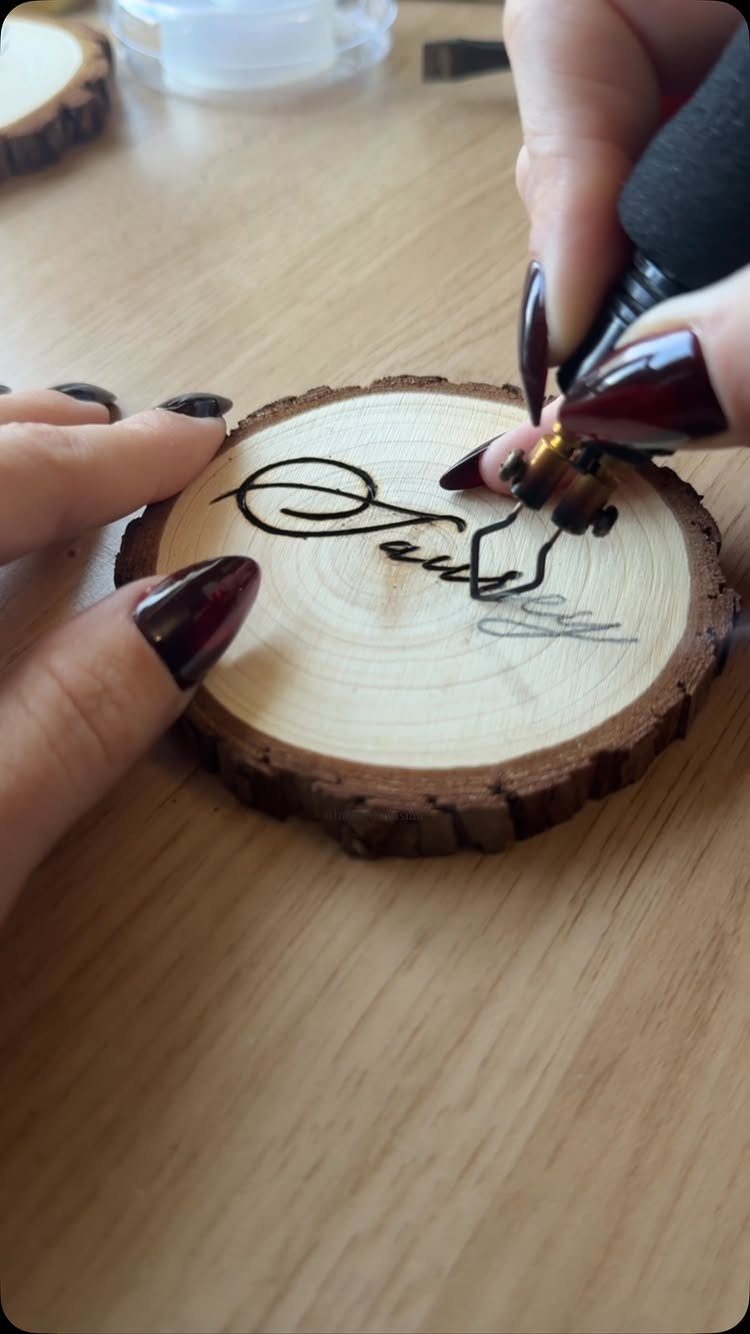Hands using a wood burning tool to create a design on a wood slice coaster.