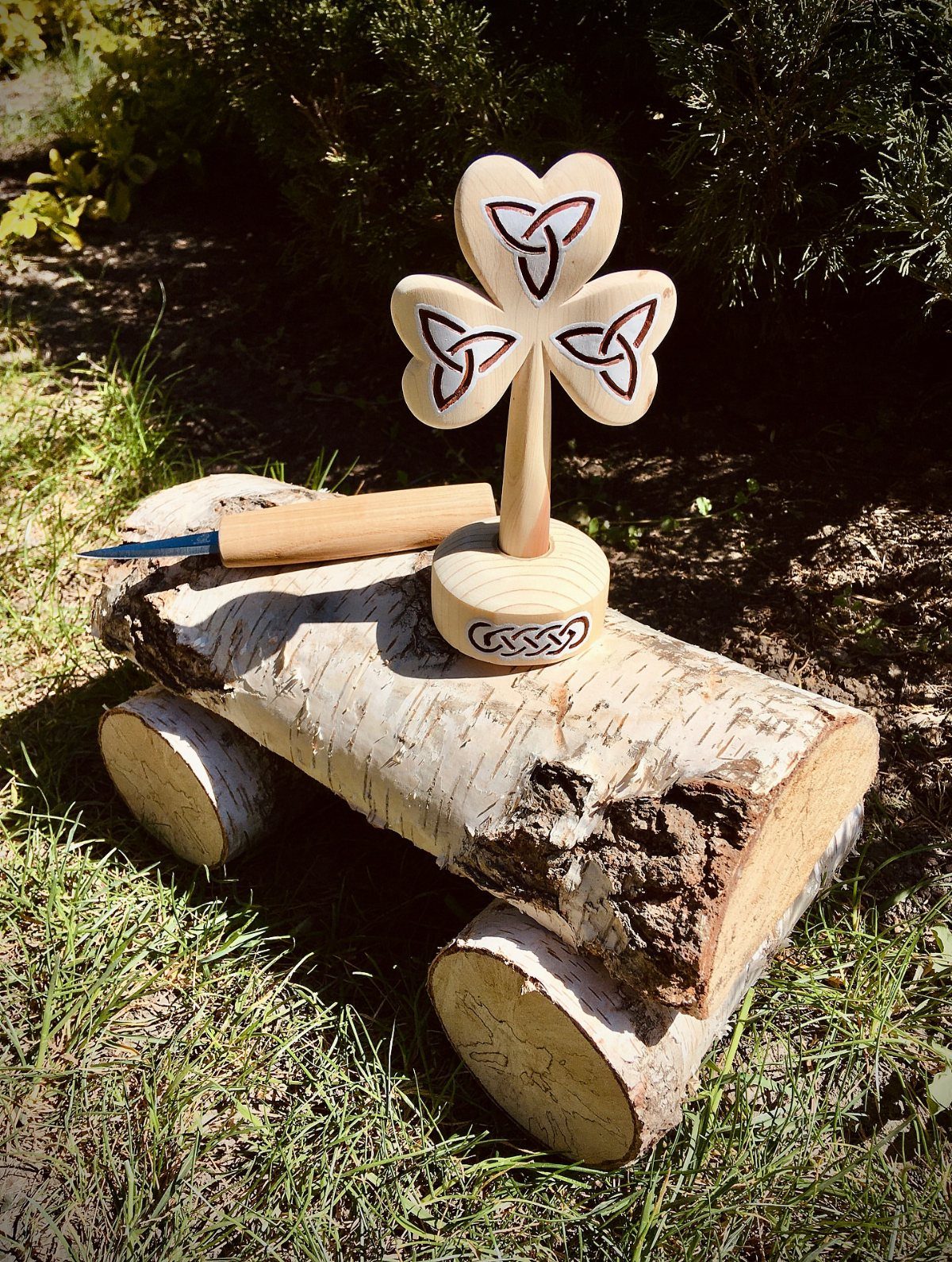Wooden shamrock with burnt Celtic knots standing on a birch log in grass.