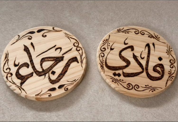Two circular wooden coasters featuring burned Arabic calligraphy and decorative floral border swirls.