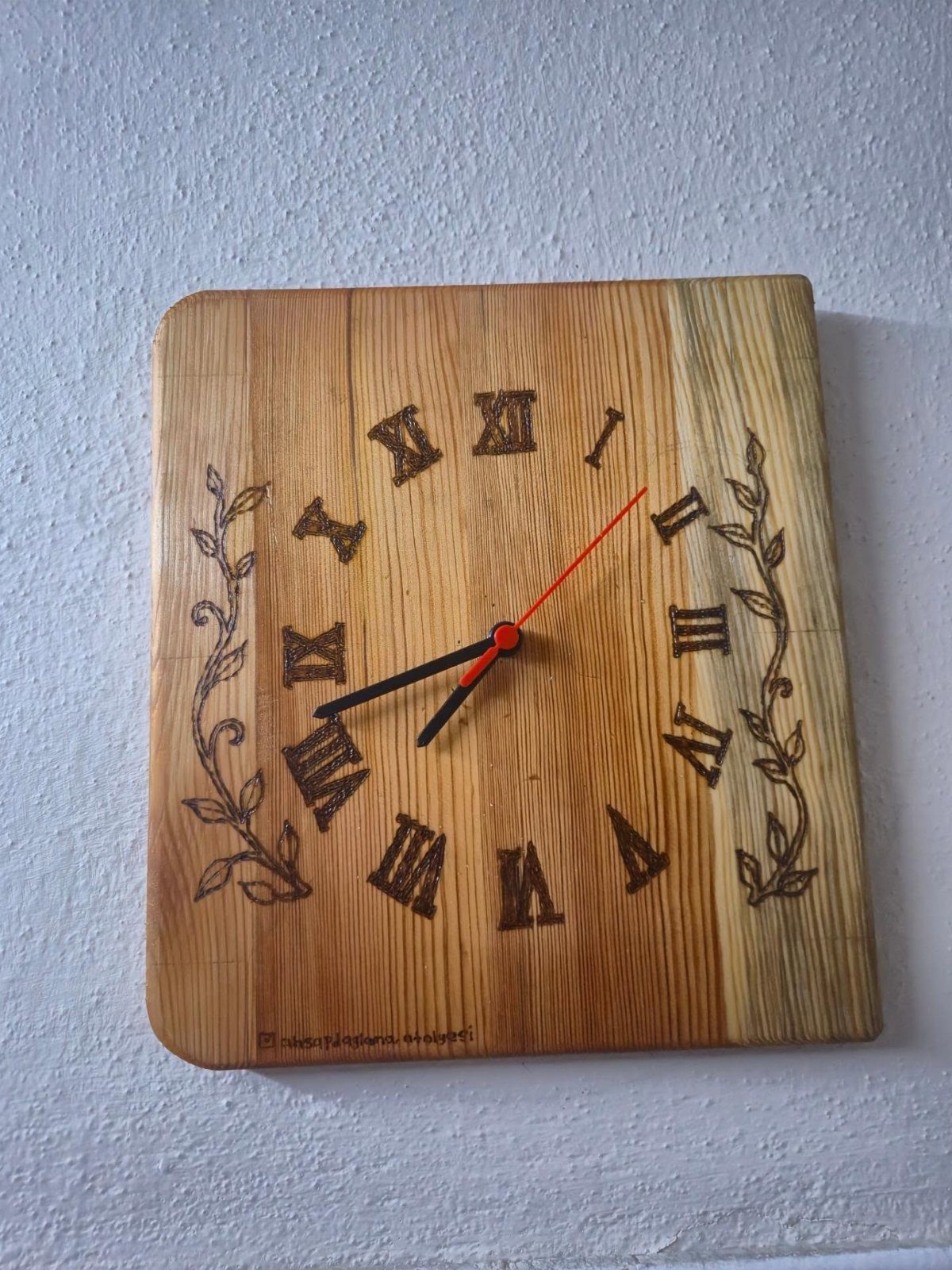 Square wooden clock with burned Roman numerals and floral designs on a white wall