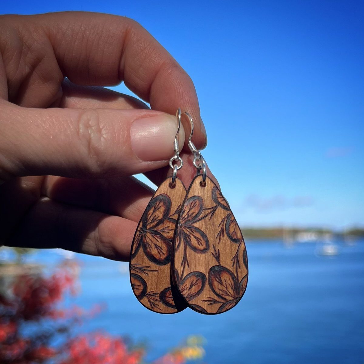 Teardrop wooden earrings with burned leaf designs held against a blue water background