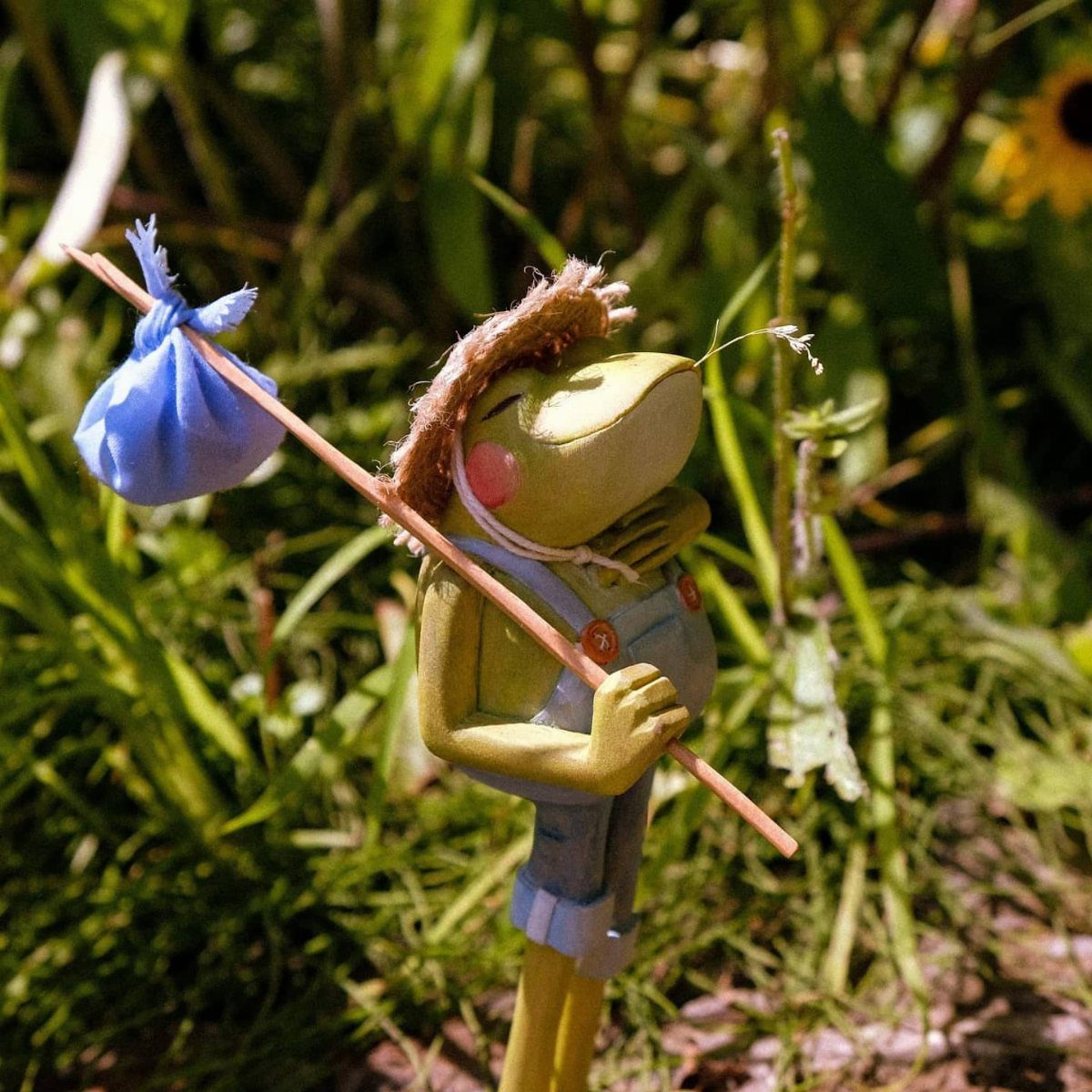 Wood Carving Animals: 14 Ideas to Try With a Knife 4 Carved wooden frog in blue overalls holding a bindle in a grassy garden