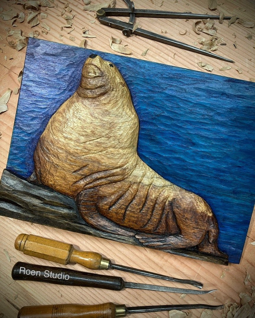 Wood Carving Animals: 14 Ideas to Try With a Knife 1 Carved wooden seal resting on a log against a blue background with carving tools.