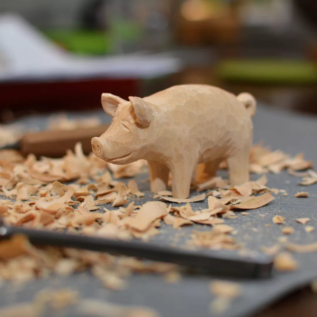 Wood Carving Animals: 14 Ideas to Try With a Knife 9 Hand-carved wooden pig figurine standing among light-colored wood shavings on a grey surface.