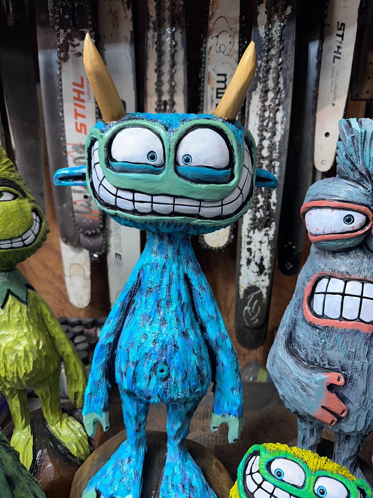 Colorful carved wooden characters with exaggerated features standing before chainsaw bars.