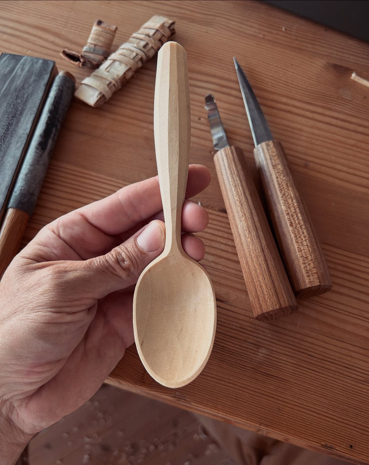 Hand-carved wooden spoon held over a workbench with carving tools and shavings