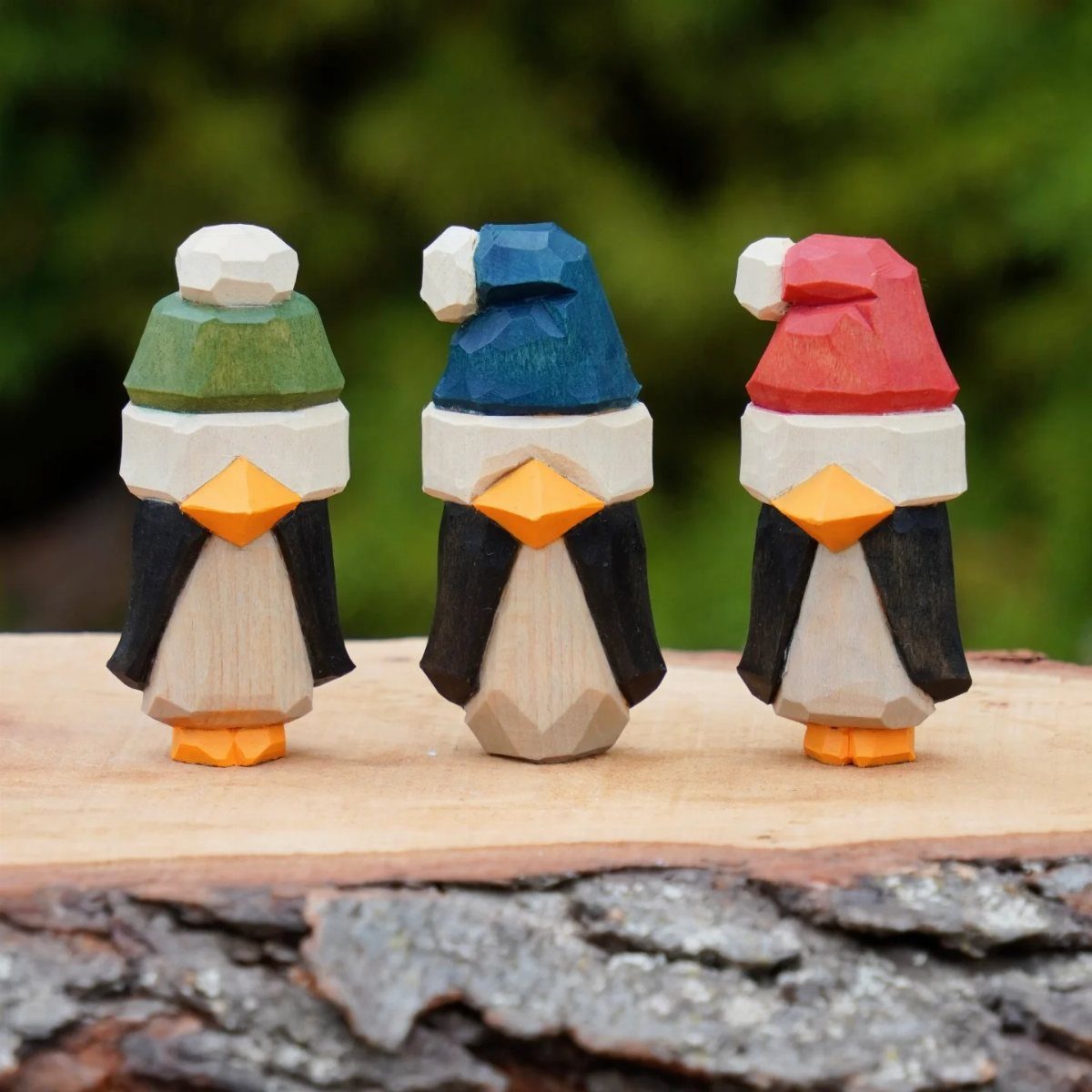 Three hand-carved wooden penguins with colorful hats standing on a rustic log slice.
