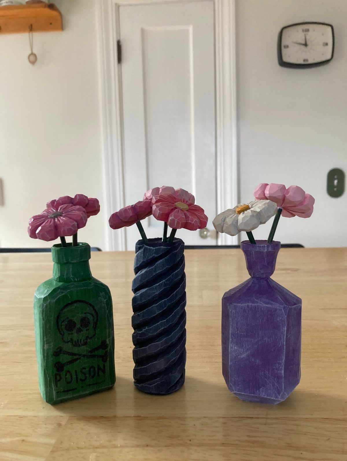 Three carved wooden vases in green, blue, and purple holding wooden flowers.