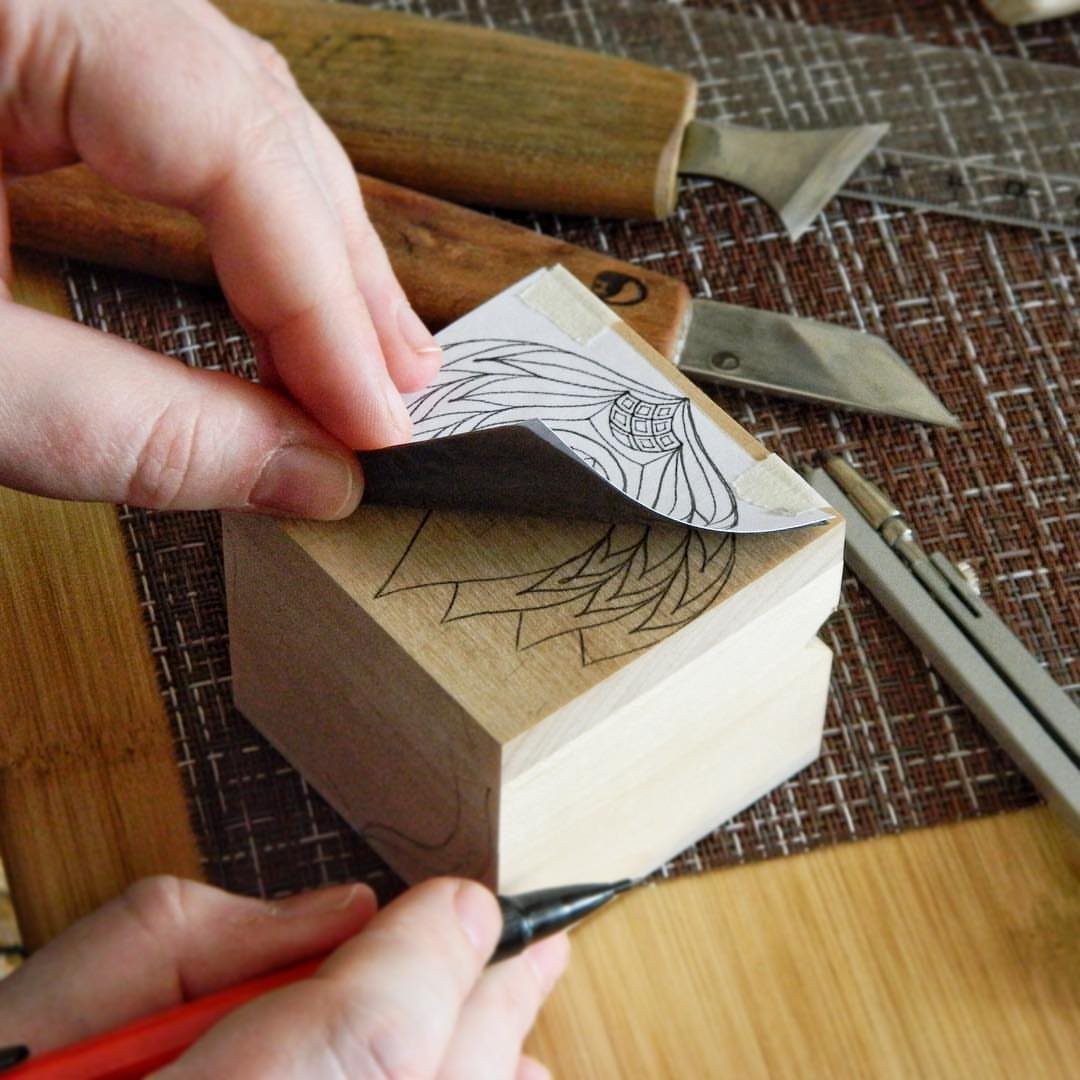 Hands lifting a paper template to show a transferred flower pattern on wood.