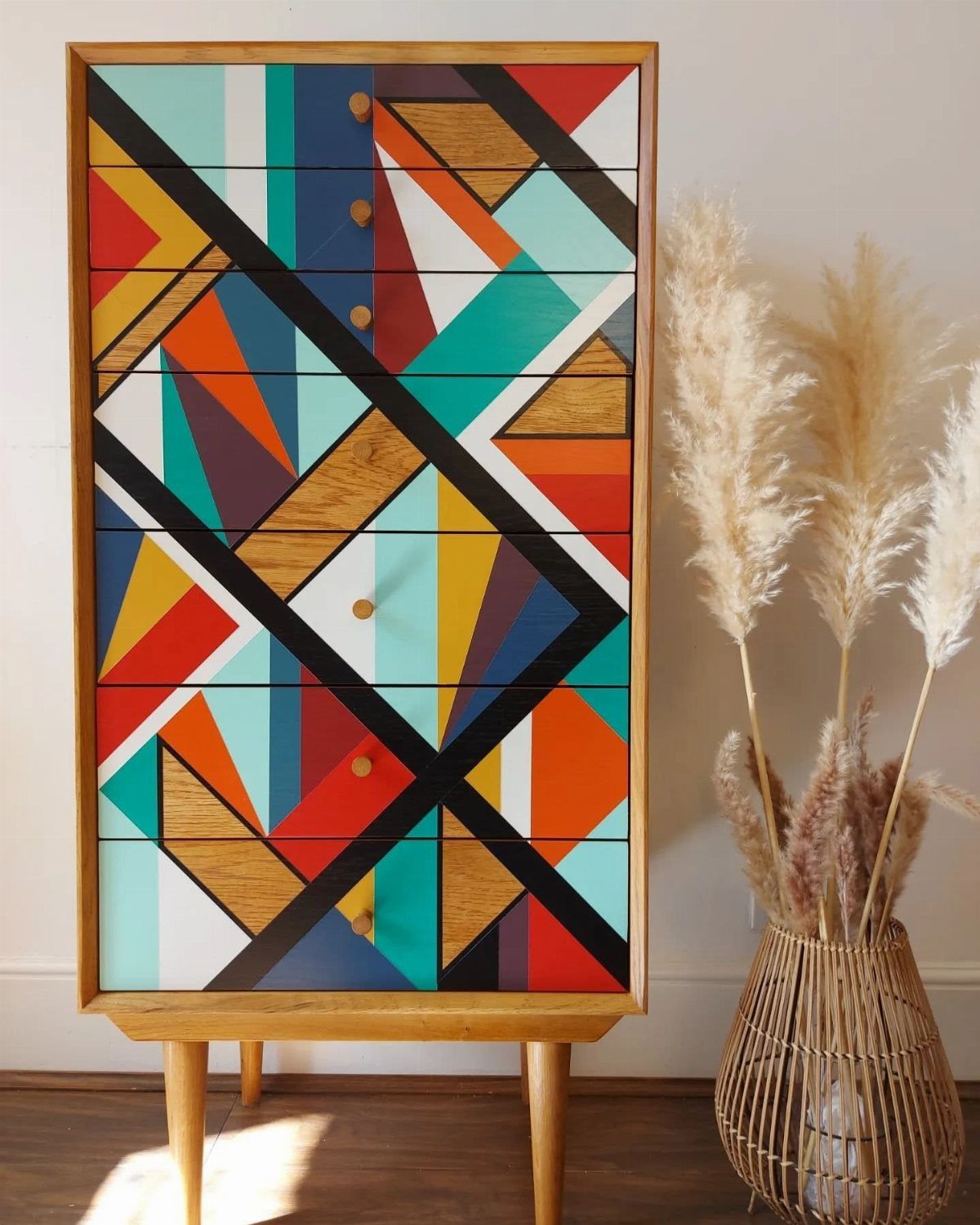Tall wooden dresser with colorful geometric patterns and tapered legs next to pampas grass.