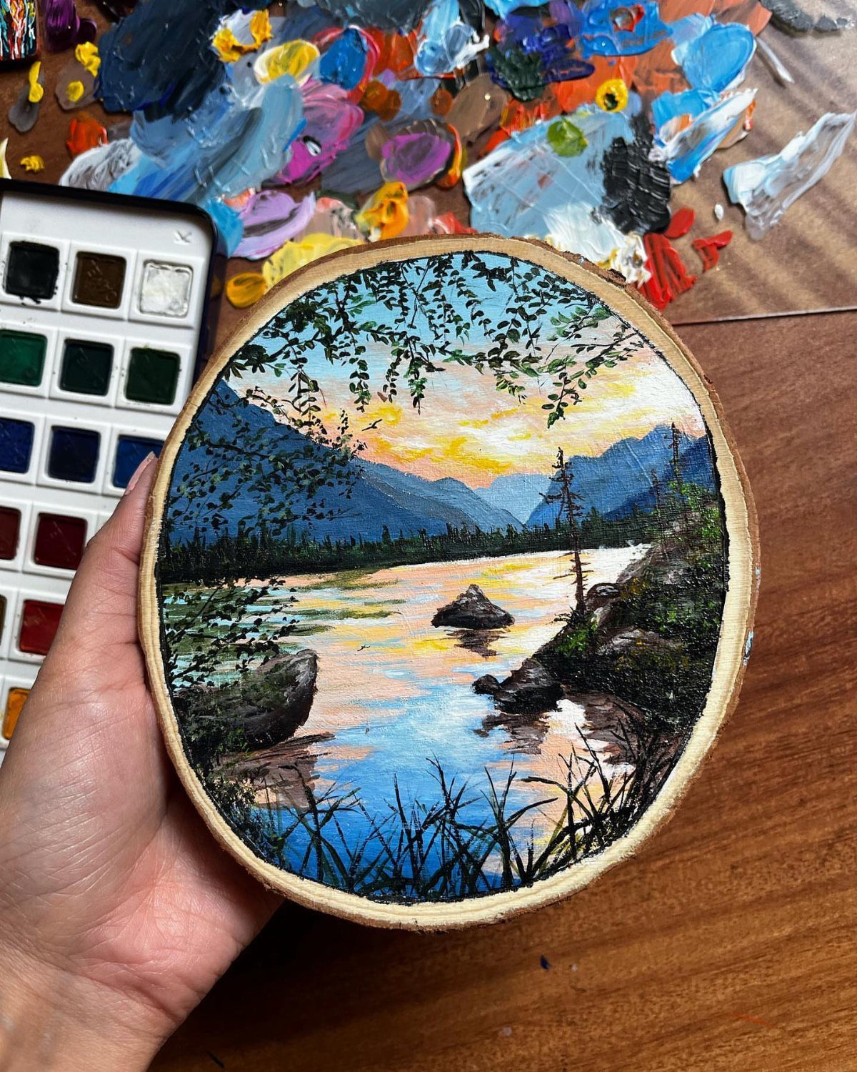 Painted wood slice showing a mountain landscape with a river at sunset.