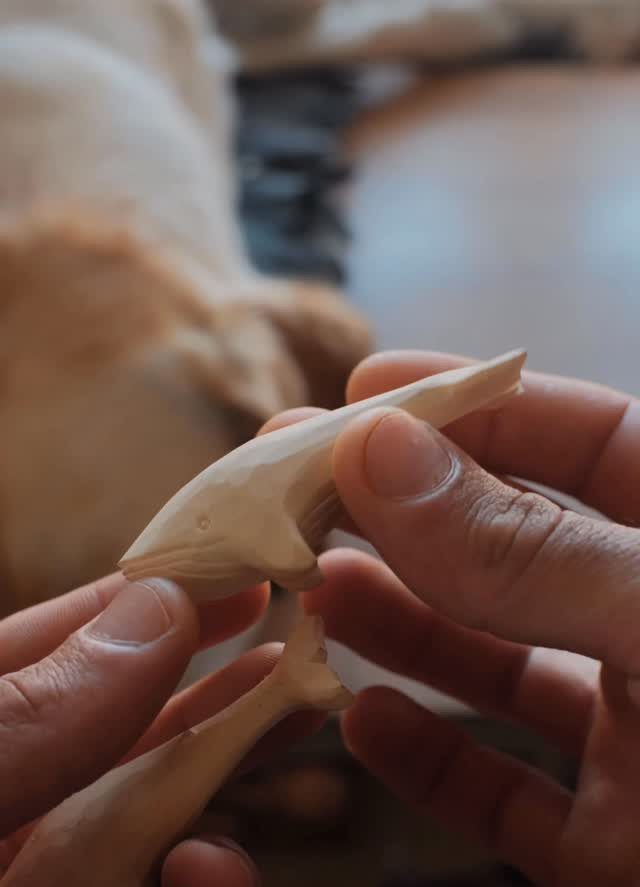 Hands holding two small hand-carved wooden whales in a home setting.