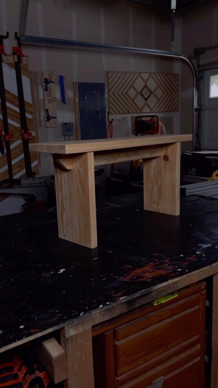 Small natural wood bench with block legs sitting on a dark workshop table.