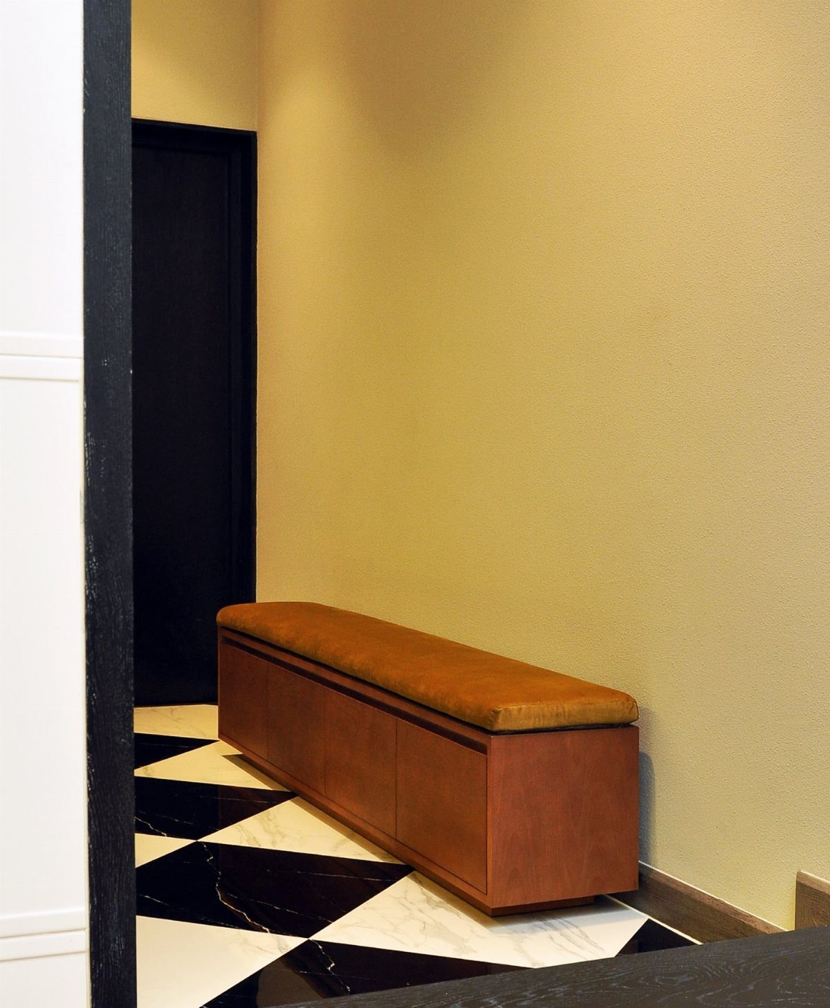 Wooden storage bench with brown upholstered cushion on a checkered marble floor.