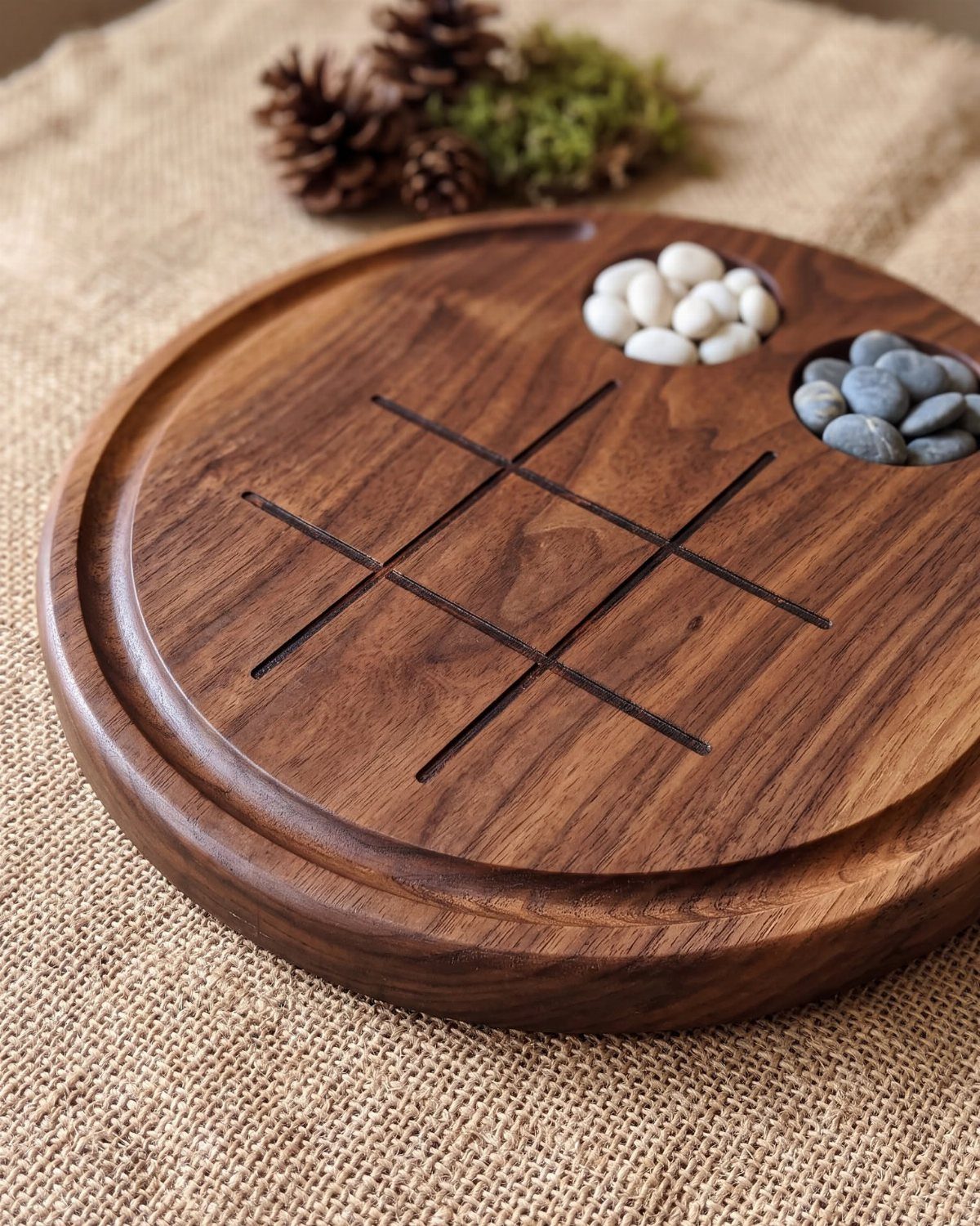 Wooden tic-tac-toe game board with white and grey pebbles on burlap fabric.