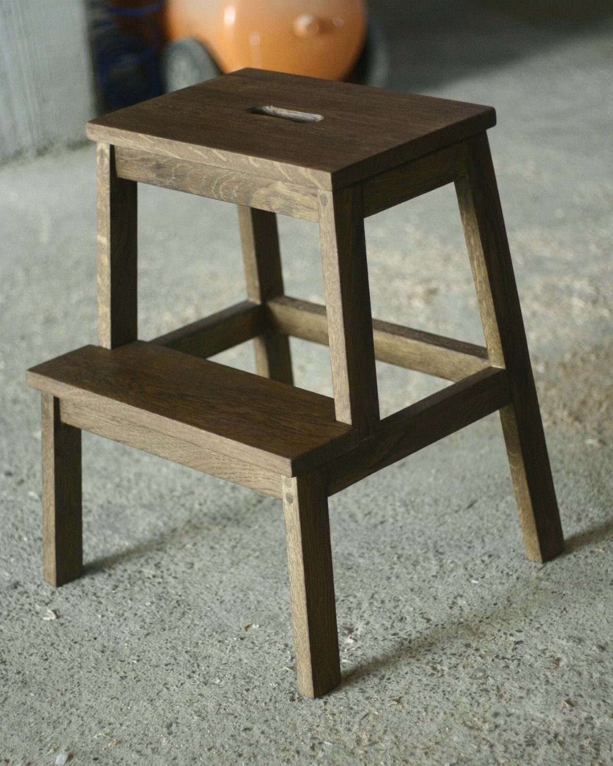 Dark brown wooden two-step stool with a handle cutout on a concrete floor.